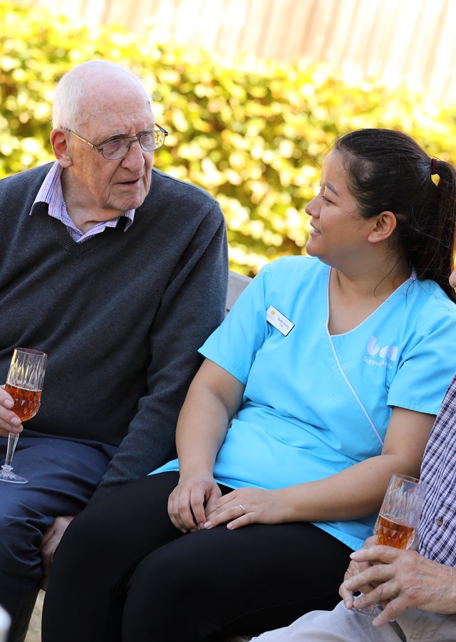 Residents are sitting on a bench in the garden, enjoying a glass of wine and chatting with a carer.