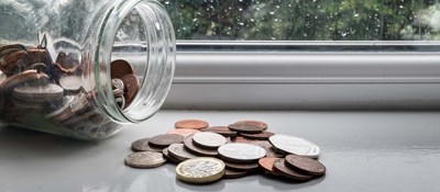 A jar full of money on its side with some of the coins spilled out on a windowsill on a rainy day