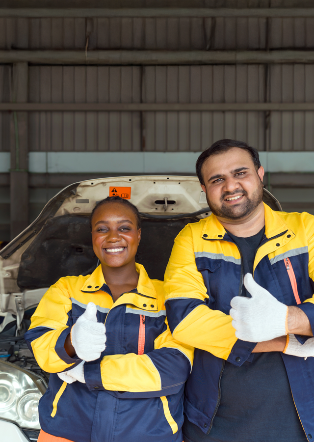 Two mechanics in work uniforms and protective gloves with thumbs up, standing in front of a white car with an open hood.