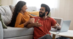 Happy couple discussing finances together on grey sofa with a laptop and coffee at home.